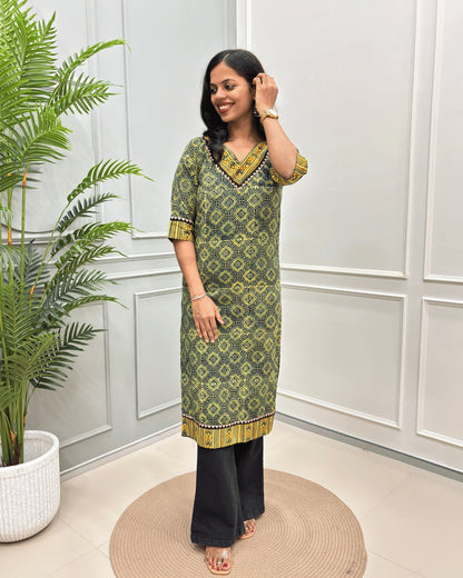 Green Women's Ajrakh Hand-block Pure Cotton Long-Kurti with Yellow and Black geometric patterns, paired with Black Flared pants, standing on a round woven mat in front of a white paneled wall with a green potted plant.