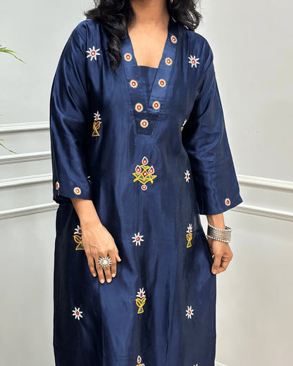 Close-up view of Navy-Blue Mashru Silk Long Kurti with colorful embroidered floral motifs and a Hand-mirror work on V-neckline, Front & Cuffs, Full Sleeves ,  standing against a light gray paneled wall. 
