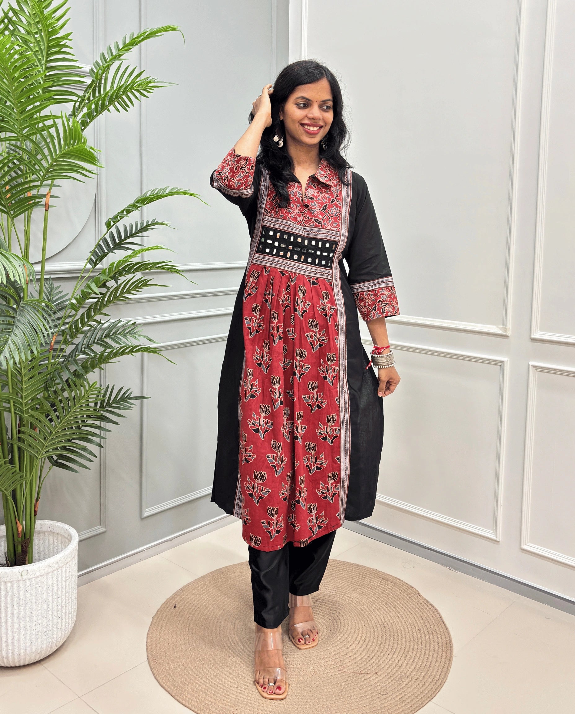 Red Women Ajrakh Vegetable Dyed Plant Based Rayon Cotton Long Kurti Featured with Blue & Black Floral print , Aline pattern with side slight, paired with black pants, standing on a round woven mat in front of a white paneled wall with a potted plant on the left.