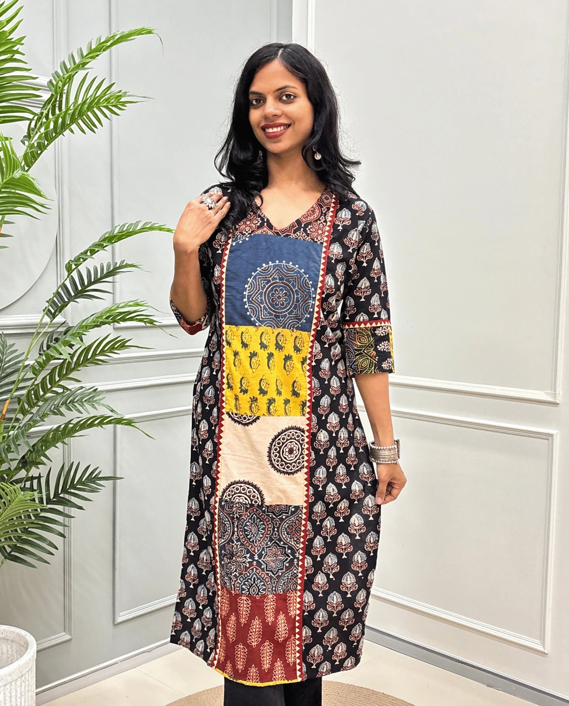 Front view of Black women's Hand-block Ajrakh pure cotton long kurti with three quarter full printed sleeves with a Black base, featuring red, yellow, beige and blue Ajrakh Geometric & badam buta print panel & cuffs, standing in front of a light gray paneled wall with a green plant on the left.