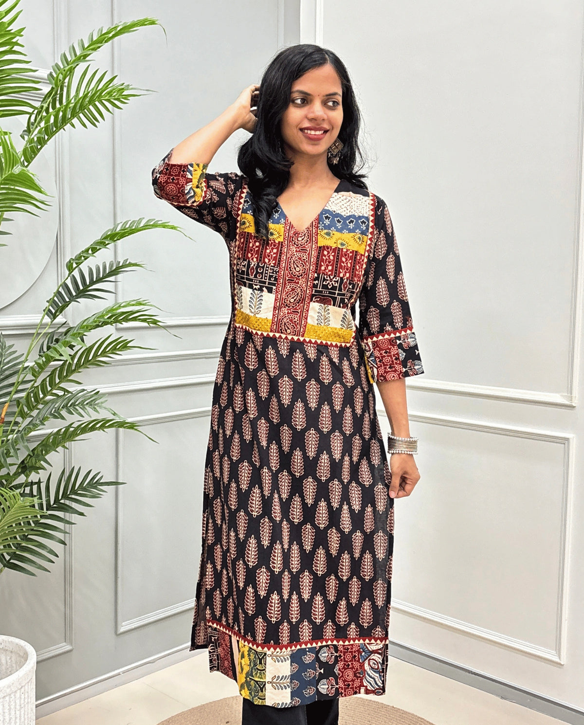 Black Women's Hand-block Ajrakh Pure cotton Long-Kurti with multicolor patchwork-style yoke on Front, Cuffs and hem, V-shaped neck, Ajrakh print on front & Sleeves, paired with black Flared pants, standing indoors against a white paneled wall next to a potted plant.