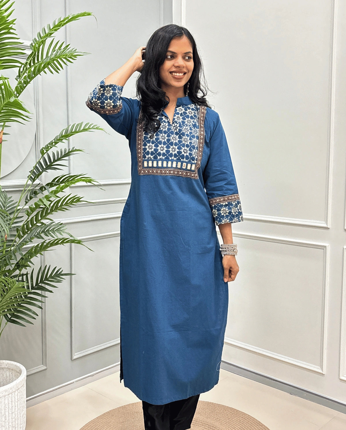 Front view of Blue Women's Hand-block Ajrakh Pure Cotton Long kurti with intricate green geometric print on the chest and sleeve cuffs, hand-mirror work on front , paired with black pants , standing on a round woven mat in front of a white paneled wall with a potted plant.