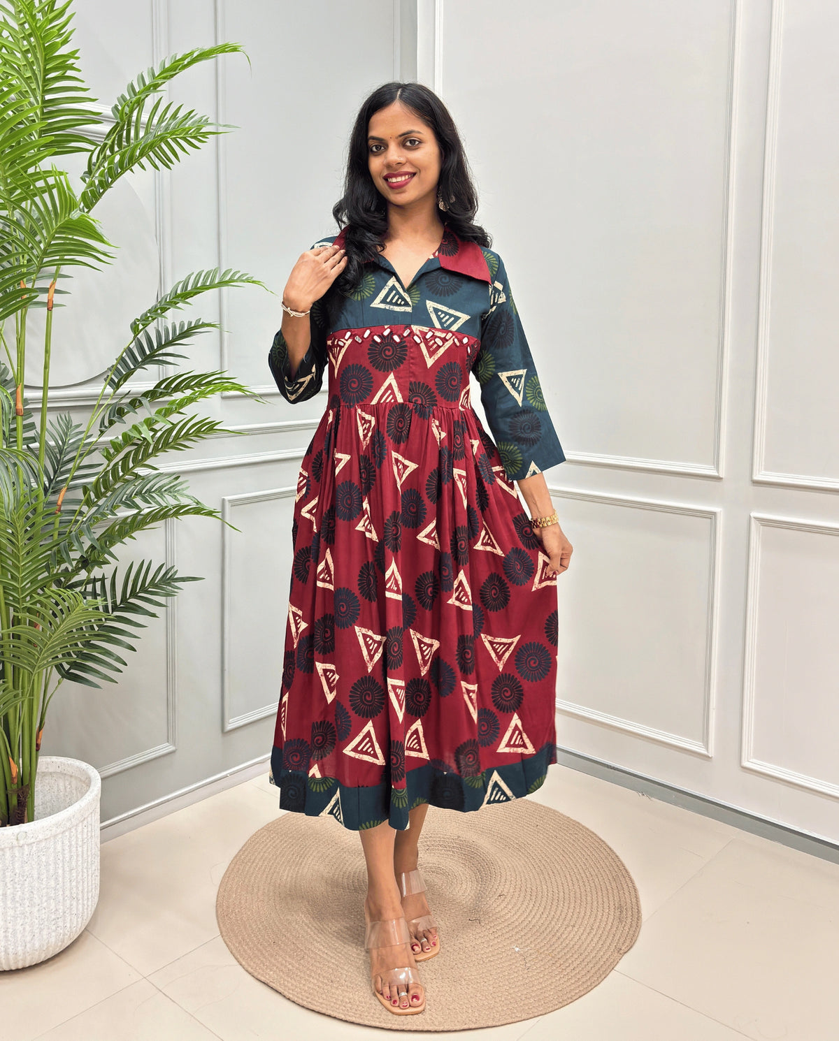 Deep Red Women's Ajrakh Vegetable dyed plant based Cotton Long-Kurti with a green on neck and cuffs, and a Red-colored dress featuring a geometric pattern of triangles and spiral in beige, black, and blue, standing on a round woven rug in front of a white paneled wall with a green potted plant on the left.