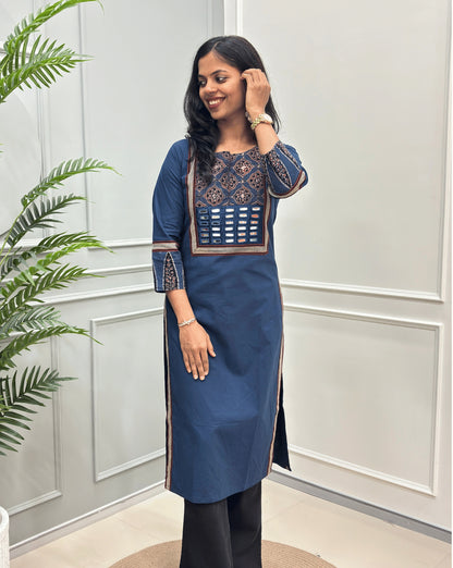 Blue Women's Hand-block Ajrakh Pure Cotton Long Kurti with a red base and abstract Black, Red & Beige  geometric embroidery on the chest and sleeve cuffs, paired with black Flared pants, standing in a decorated room with a potted plant.
