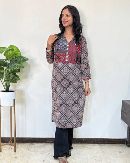 Black Women's Ajrakh Vegetable Dyed plant based cotton long kurti with multi-colour patterns, paired with black flared pants, standing in a room with a white wall and a potted plant.