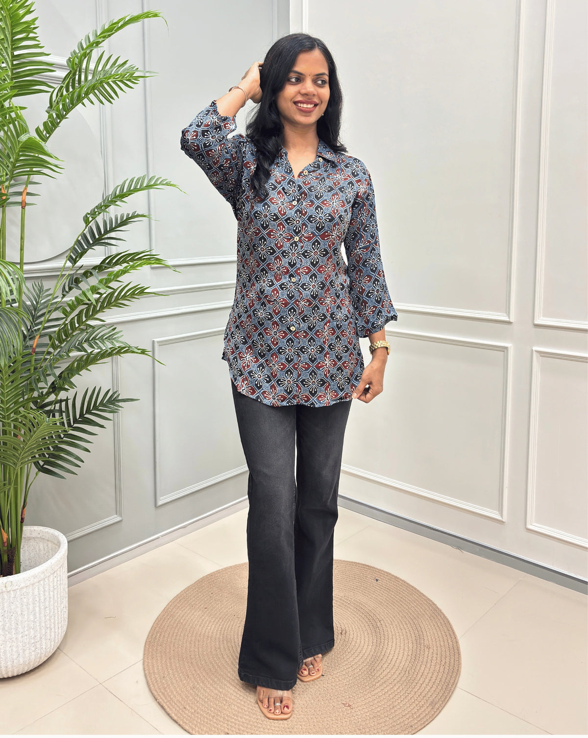 Deep Blue Women's Ajrakh Pure Modal Silk Shirt with a Floral print , black flared pants, standing on a round woven rug in front of a white paneled wall with a potted plant on the left.