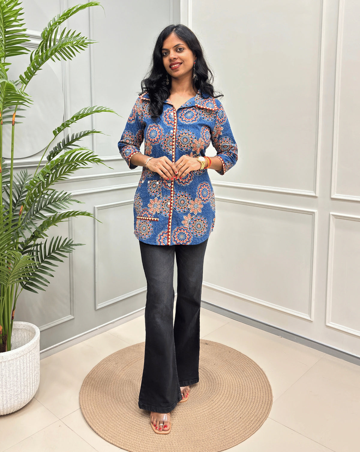 Blue women's Ajrakh pure cotton Shirt with a blue base and Multi color floral print,  paired with black flared pants. Standing on a round woven rug against a light paneled wall with a potted plant at left.