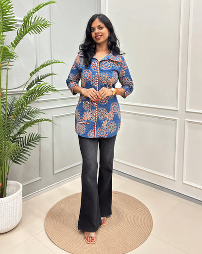 Blue women's Ajrakh pure cotton Shirt with a blue base and Multi color floral print,  paired with black flared pants. Standing on a round woven rug against a light paneled wall with a potted plant at left.