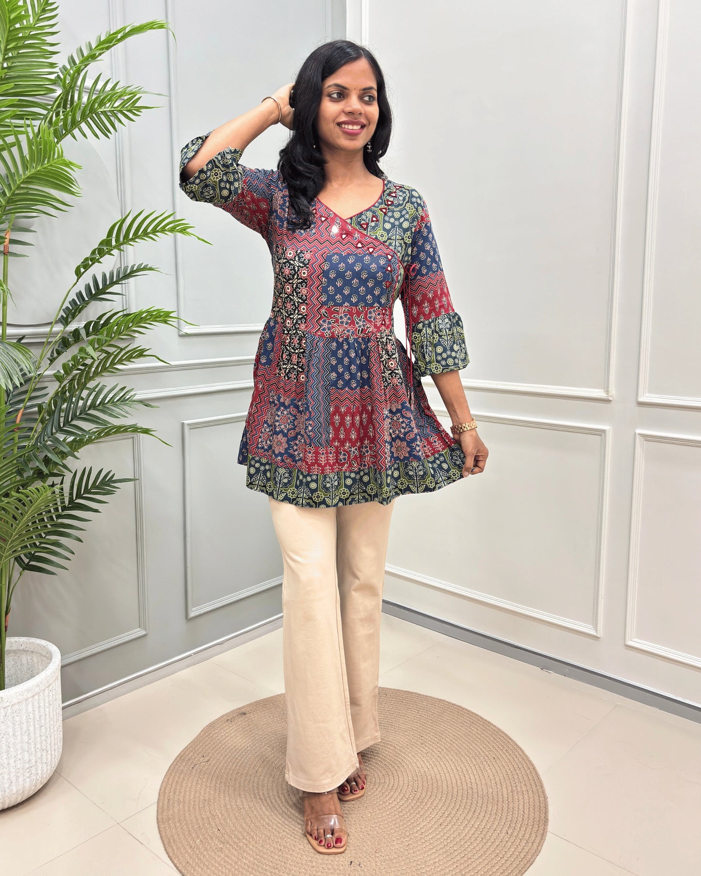 Multi Women's Ajrakh Vegetable Dyed plant based cotton Short Kurti with colourful  geometric pattern, paired with beige flared pants, standing on a round woven mat in front of a white paneled wall with a potted plant.
