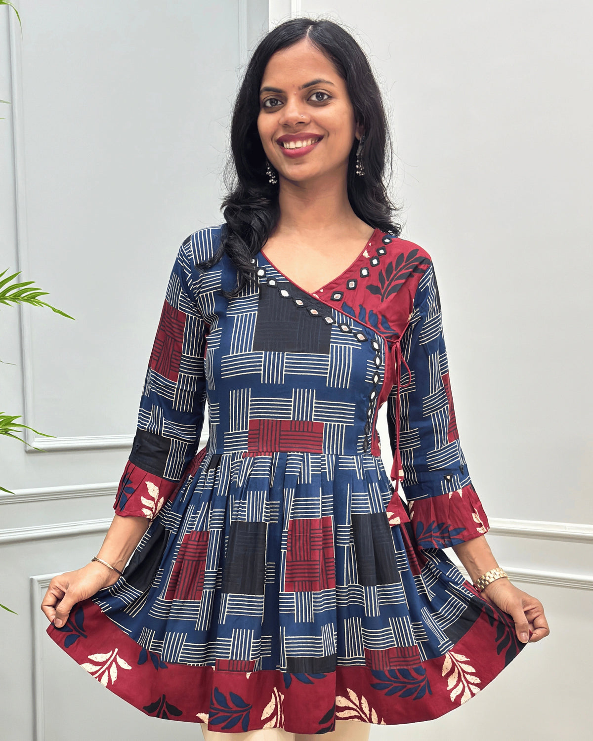 Close-up of Blue women's Ajrakh plant based cotton Short kurti with a colorful geometric and circular print, Hand-Mirror work on V- neckline along with lace, Leaf print on red base on half neck, bottom, & sleeves cuffs, paired with beige pants, standing in front of a white paneled wall with a green plant on the left.