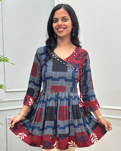 Close-up of Blue women's Ajrakh plant based cotton Short kurti with a colorful geometric and circular print, Hand-Mirror work on V- neckline along with lace, Leaf print on red base on half neck, bottom, & sleeves cuffs, paired with beige pants, standing in front of a white paneled wall with a green plant on the left.