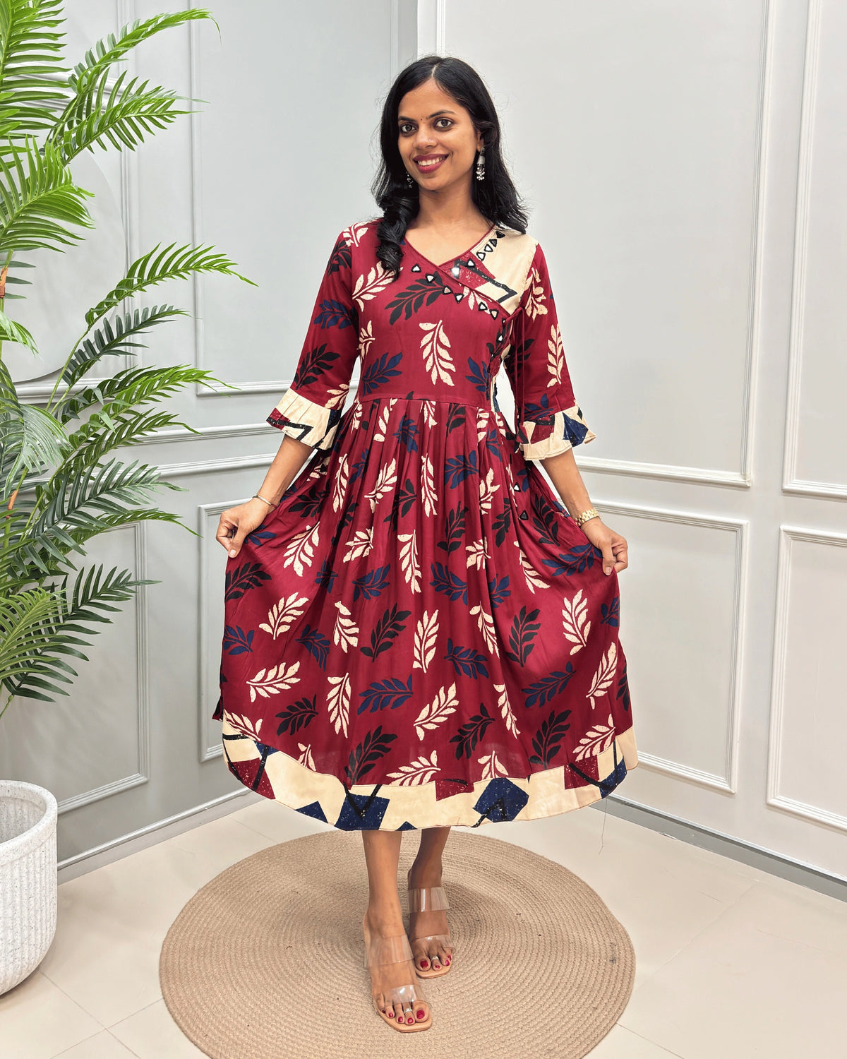 Red Women's Vegetable dyed Plant based Cotton Long Kurti with three-quarter Frill sleeve , a red base and cream and navy blue leaf patterns, standing on a round woven mat in front of a white paneled wall with a potted plant.