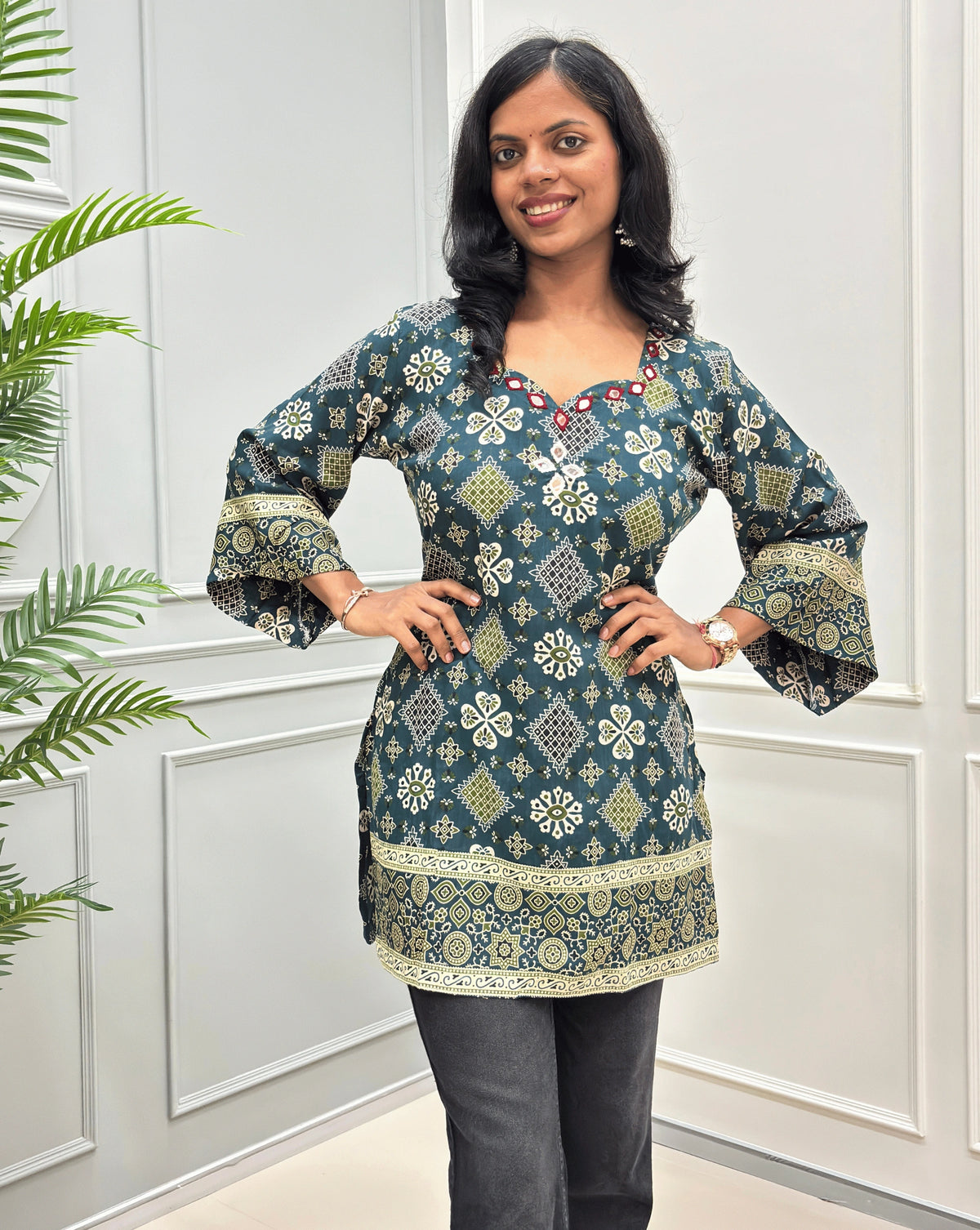 A green & beige printed cotton short kurti with beautiful bell sleeves, standing in front of a white paneled wall.