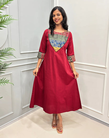Front view of Red women's Ajrakh Vegetable dyed plant based cotton long kurti with a heavily embroidered multicolor Mashru yoke on front chest & sleeves cuffs along with  pocket, with black piepin, standing in Front of white paneled room , on left side plotted plant .