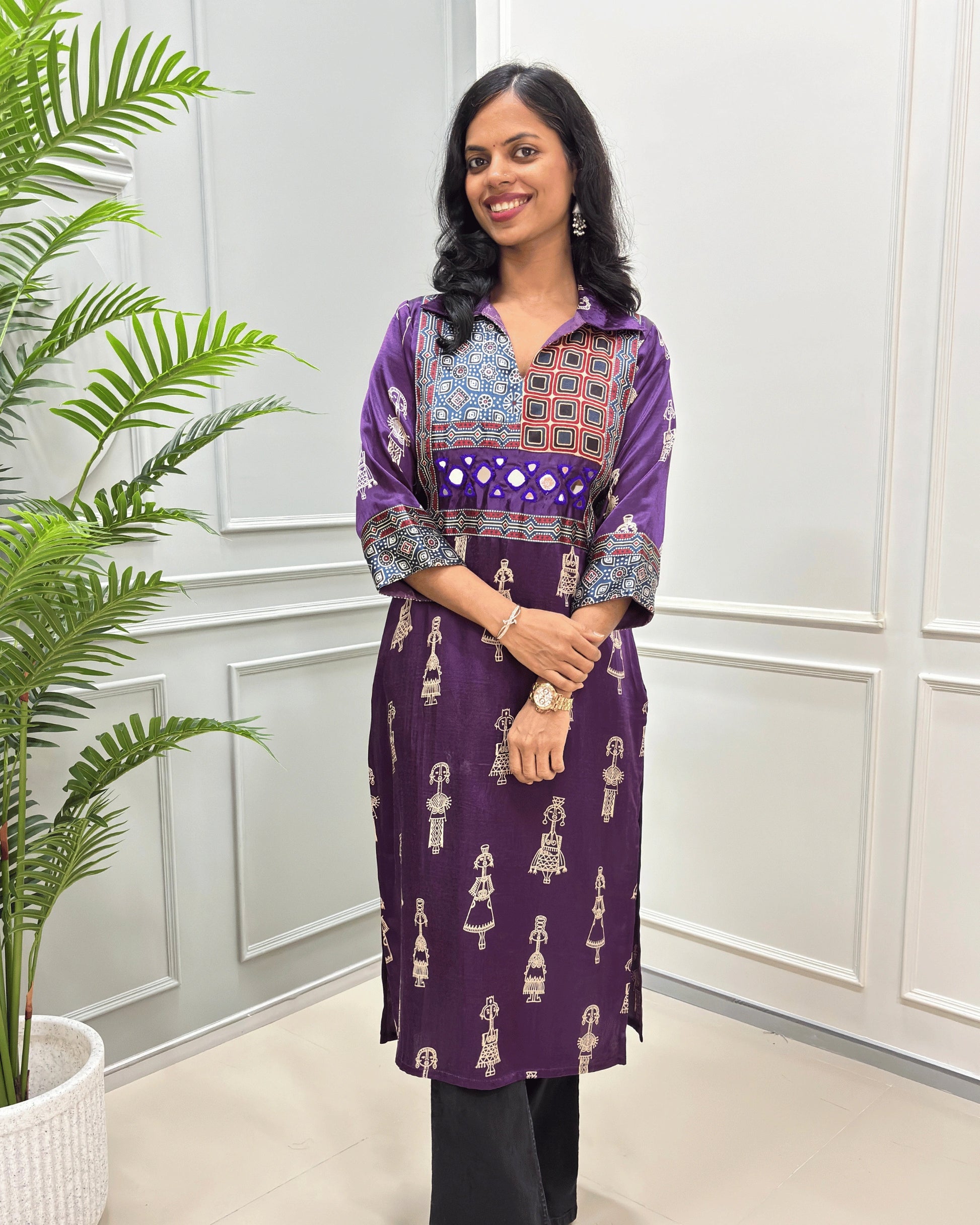 Close-up of Purple Ajrakh Mashru silk Long kurti with multicolor geometric , warli print on front & sleeves along eith hand -mirror work on front , Collar V shaped neck patterns, standing in front of a white paneled wall with a green plant on the left.