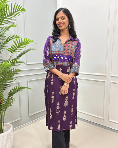 Close-up of Purple Ajrakh Mashru silk Long kurti with multicolor geometric , warli print on front & sleeves along eith hand -mirror work on front , Collar V shaped neck patterns, standing in front of a white paneled wall with a green plant on the left.