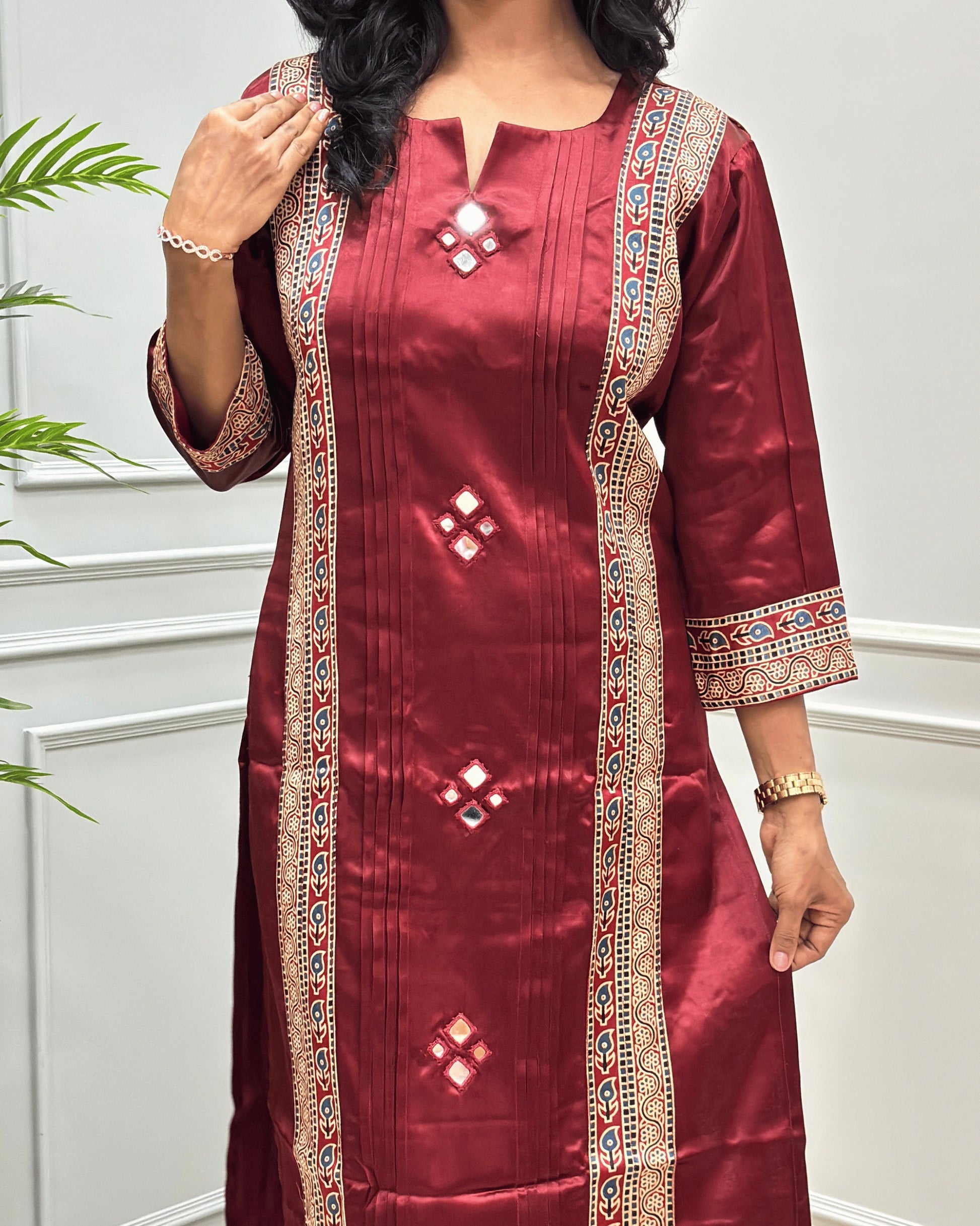 Close-up of Deep Red of Ajrakh Mashru Silk Long-kurti with intricate beige, red, and blue patterned borders and mirror work, Round V-neck,  shown in a lifestyle setting against a white paneled wall.