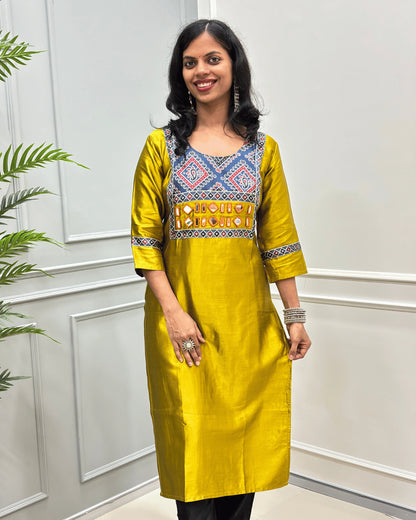 Front view of Mustard Yellow Ajrakh Mashru Silk Long-kurti with a blue, red, and white in Bandhani print , featuring Hand-mirror work with Yellow thread work embellishments , blue & red patterned cuffs, standing against a white paneled wall.