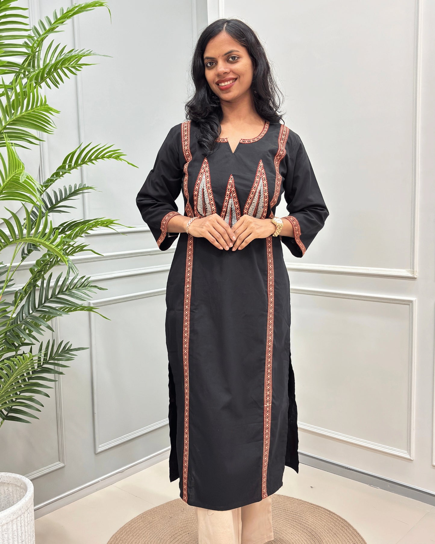 Front-view of Black Women's Hand-block Ajrakh Pure Cotton Long Kurti with Red and Black embroidered details on the V-neckline and borders, standing in a decorated room with a potted plant.