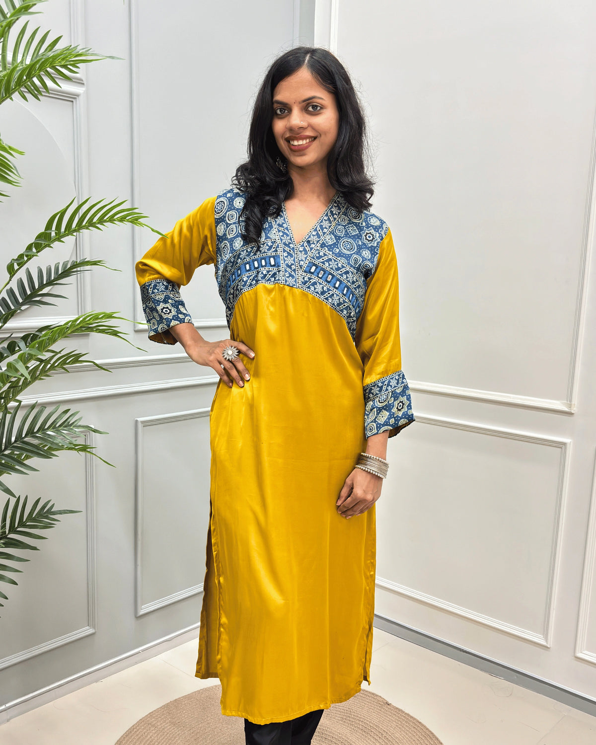 Front view of Mustard-Yellow women's Ajrakh Modal Silk Long kurti featured with Blue base & ajrakh print with white on neck & cuffs , hand-mirror work on chest standing in a white paneled room.