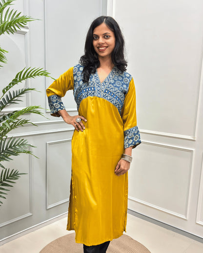 Front view of Mustard-Yellow women's Ajrakh Modal Silk Long kurti featured with Blue base & ajrakh print with white on neck & cuffs , hand-mirror work on chest standing in a white paneled room.