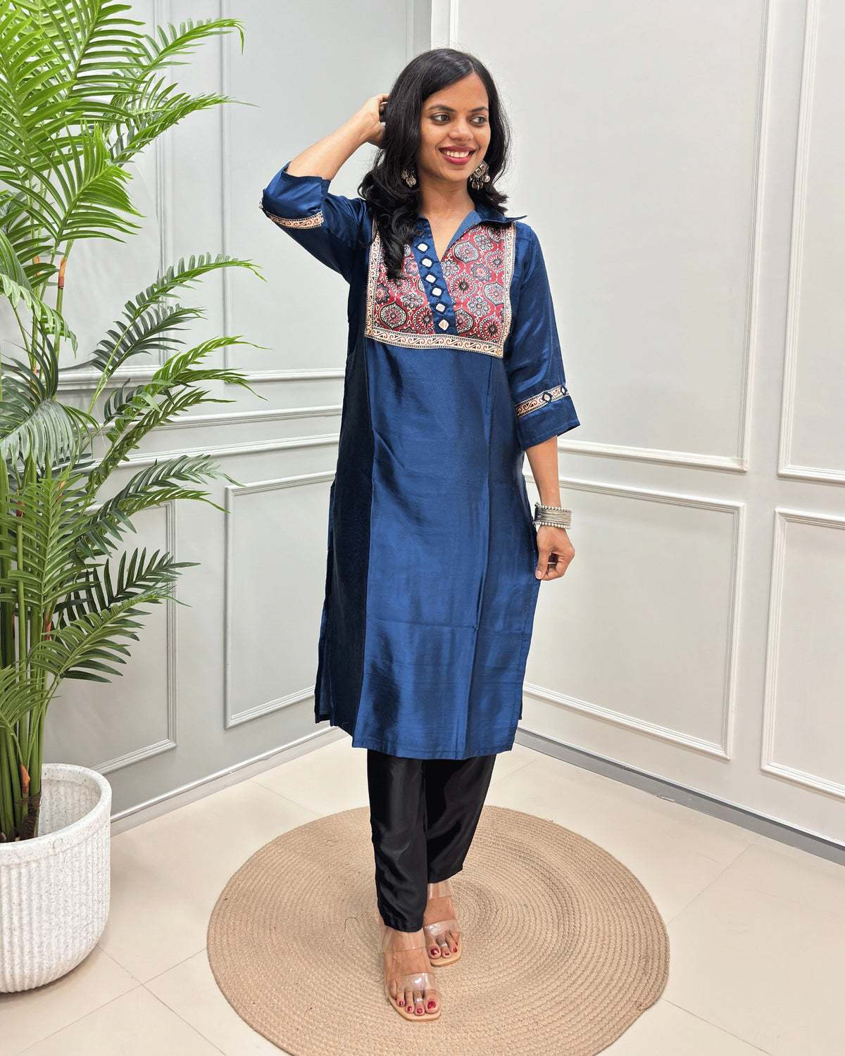 Blue Women's Ajrakh Mashru Silk Long Kurti with colorful embroidered detailing on the chest and sleeves, paired with black pants, standing in a decorated room with a potted plant and paneled wall.