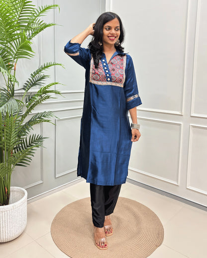 Blue Women's Ajrakh Mashru Silk Long Kurti with colorful embroidered detailing on the chest and sleeves, paired with black pants, standing in a decorated room with a potted plant and paneled wall.
