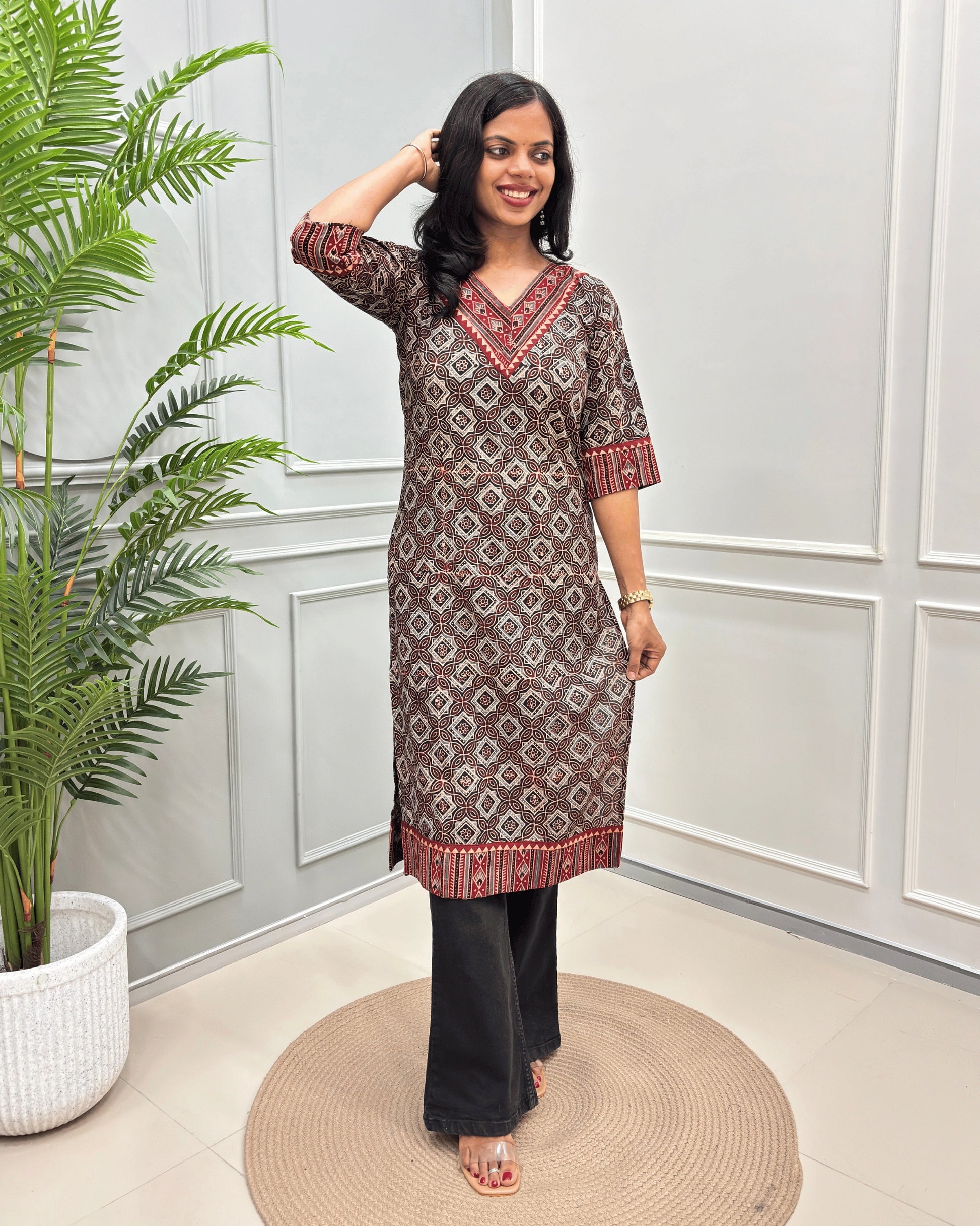 Black Women's Ajrakh Hand-block Pure Cotton Long-Kurti with red and Beige geometric patterns, paired with Black Flared pants, standing on a round woven mat in front of a white paneled wall with a green potted plant.