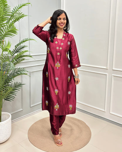 Maroon women's Pure Mashru Silk Long Kurti with yellow and white floral embroidery, & Ahir work on front & Sleeves cuffs , paired with maroon pants, standing on a round woven mat in front of a white paneled wall with a green plant to the side.