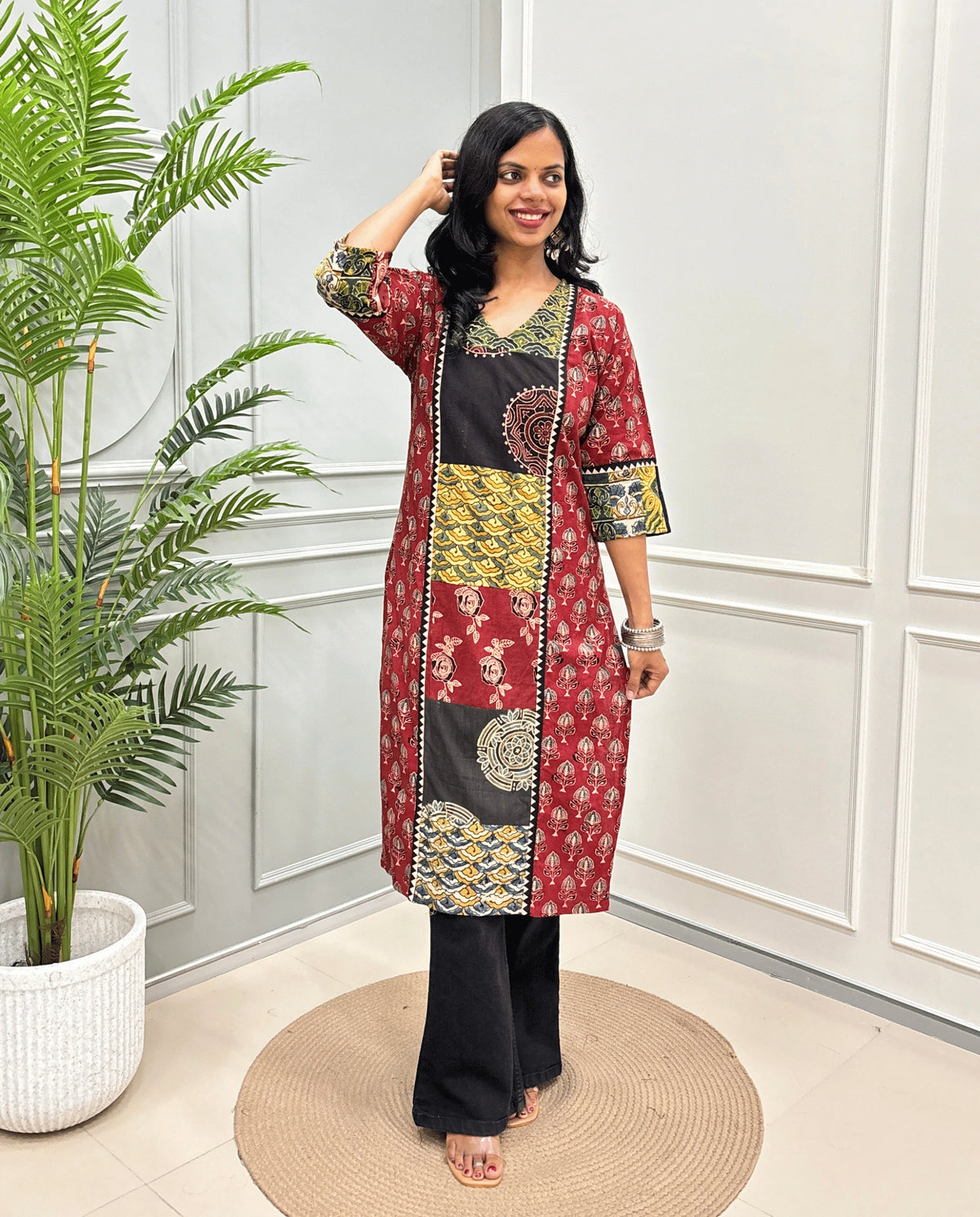 Red women's Hand-block Ajrakh  pure cotton Long Kurti with three-quarter sleeves, Ajrakh print on front and sleeves and black wide-leg pants, standing on a round woven mat in front of a white paneled wall next to a potted palm.
