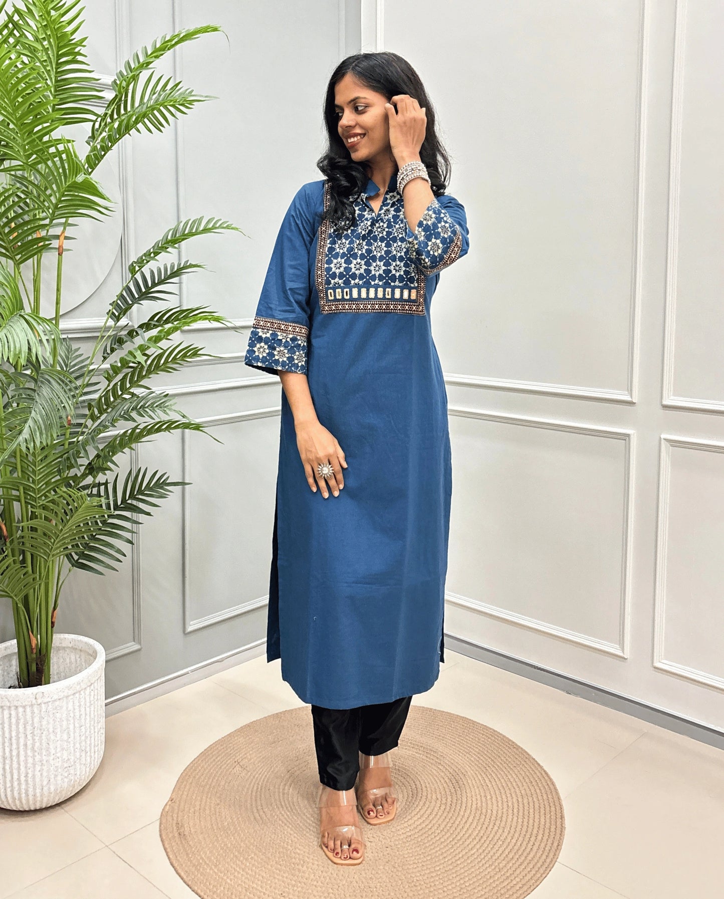 Blue Women's Hand-block Ajrakh Pure Cotton Long kurti with intricate white geometric print on the chest and sleeve cuffs, paired with black pants , standing on a round woven mat in front of a white paneled wall with a potted plant.