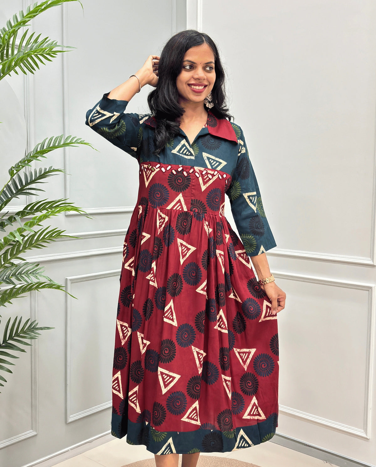 Deep Red Women's Ajrakh Vegetable dyed plant based Cotton Long-Kurti with a green neck and cuffs, and a Red-colored collar & dress featuring a geometric pattern of triangles and spiral in beige, black, and blue, Hand-mirror work on front, standing on a round woven rug in front of a white paneled wall with a green potted plant on the left.