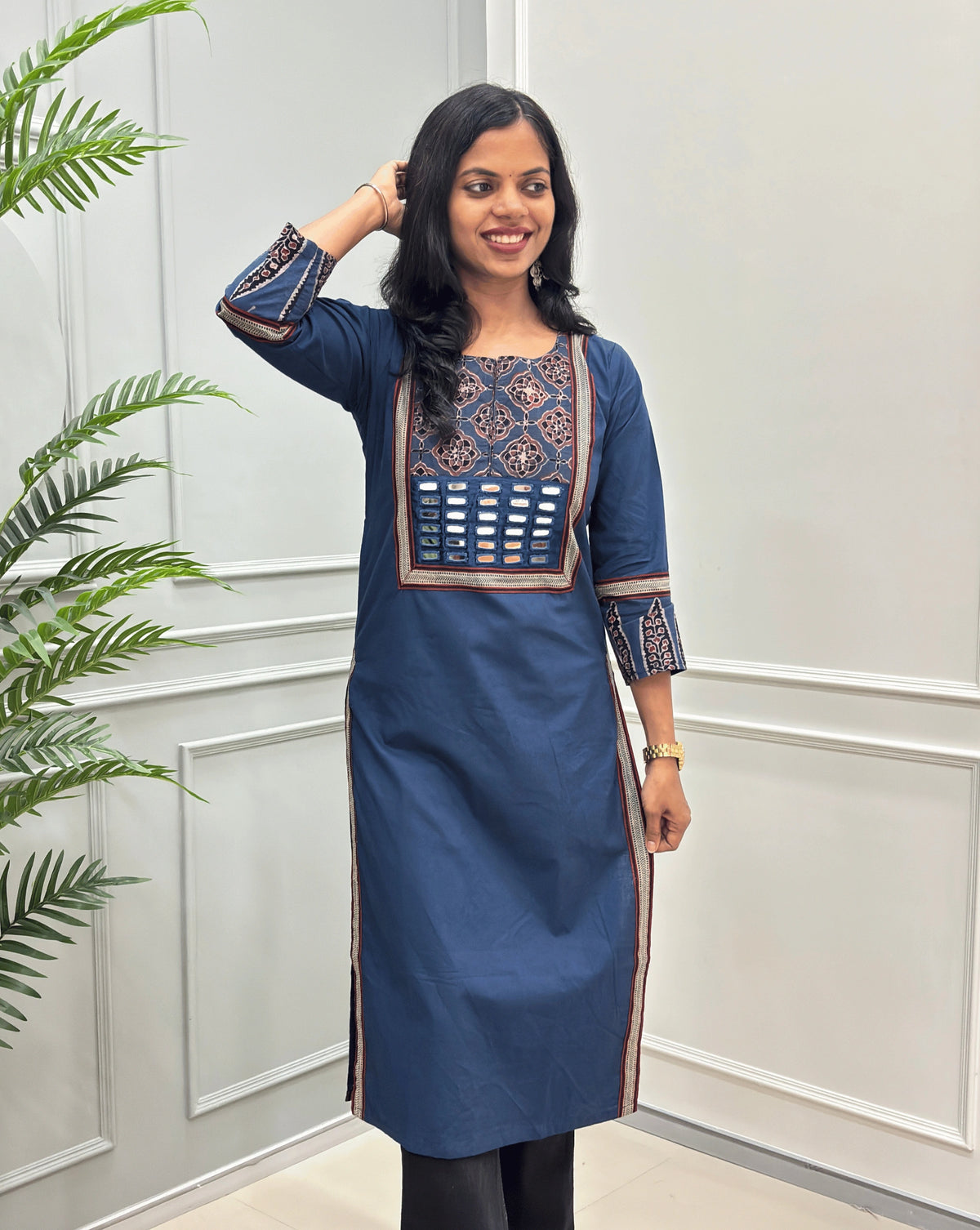 Front View of Blue Women's Hand-block Ajrakh Pure Cotton Long Kurti with a red base and abstract Black, Red & Beige geometric embroidery on the chest and sleeve cuffs, Hand-mirror work on Front, Ajrakh print border on Sides & Front  paired with black Flared pants, standing in a decorated room with a potted plant.