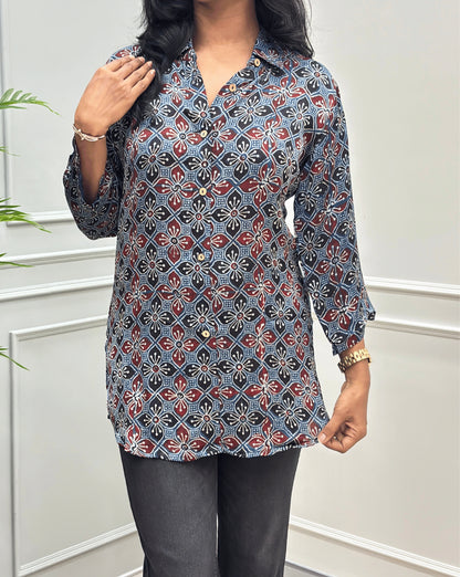 Close-up of Blue Women's Ajrakh Pure Modal Silk Shirt with Full-Sleeves, with a Floral print on blue, red, and white geometric pattern, black flared pants, standing on a round woven rug in front of a white paneled wall with a potted plant on the left