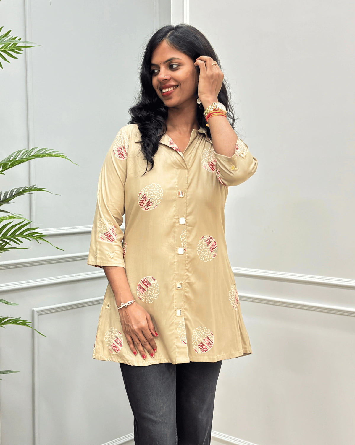 Front view of Beige Women's Ajrakh Vegetable dyed Plant based A-line cotton Short kurti with pink & white floral embroidery, Hand-mirror work on front look a like buttons , Sleeves Cuffs & pockets, paired with black flared jeans, standing on a round woven rug in a modern room with a potted plant.