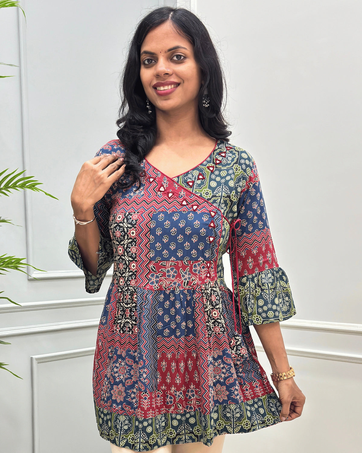 Close-up of Multi colour women's Ajrakh Plant based cotton short-kurti with a multicolor patchwork print featuring navy blue, red, green, and beige geometric and floral patterns, Angrakha Pattern with Hand-mirror work on V-shaped Neckline along with Lace, set against a light gray paneled wall.