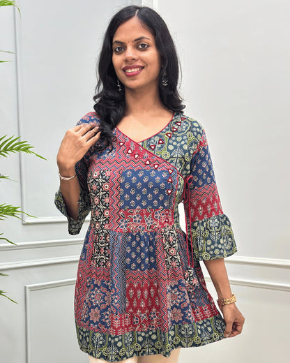 Close-up of Multi colour women's Ajrakh Plant based cotton short-kurti with a multicolor patchwork print featuring navy blue, red, green, and beige geometric and floral patterns, Angrakha Pattern with Hand-mirror work on V-shaped Neckline along with Lace, set against a light gray paneled wall.