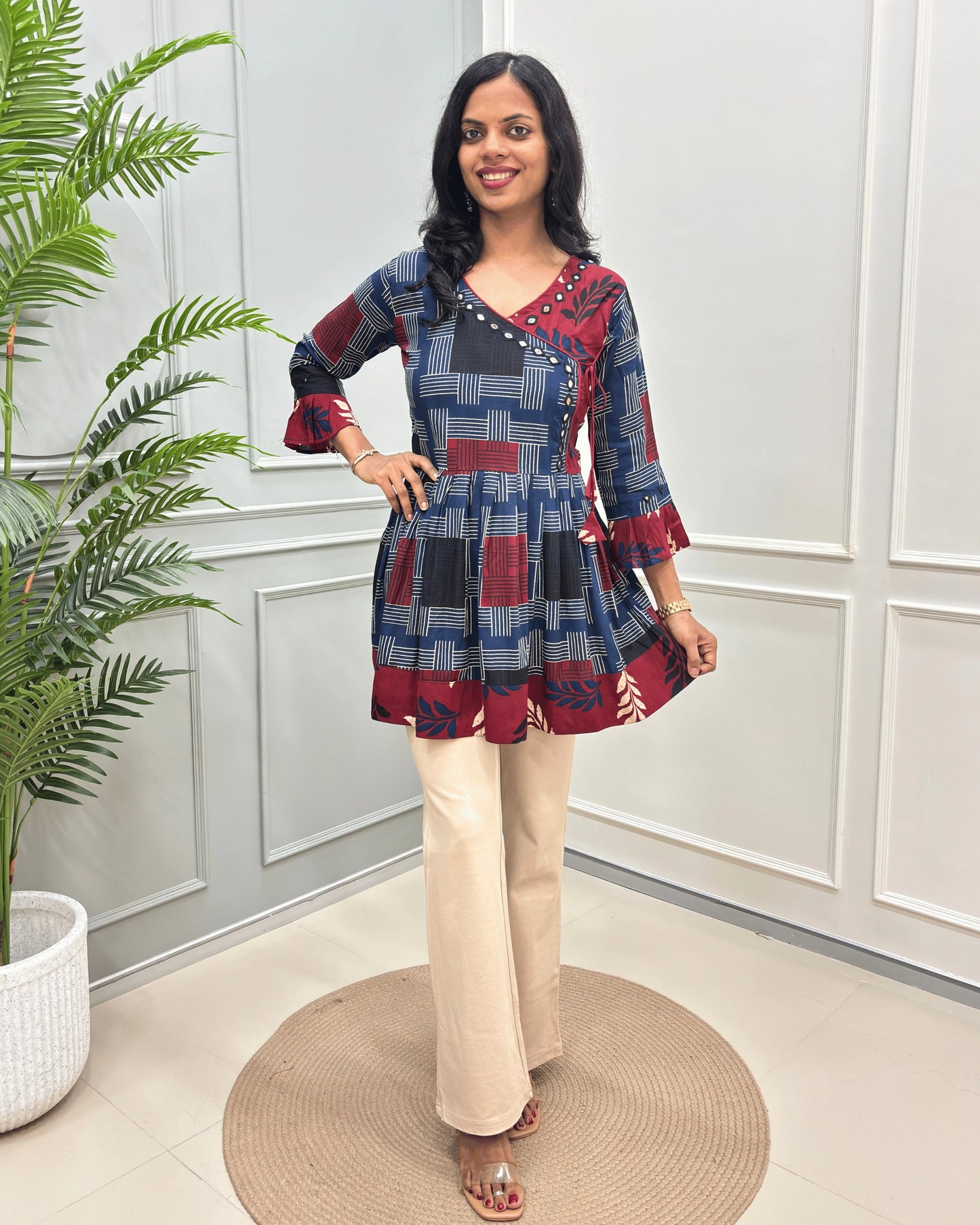 Blue Women's Ajrakh Vegetable dyed plant based Cotton Short-Kurti with three-quarter frill sleeves, and waist to bottom,  paired with beige flared pant , standing on a round woven rug in front of a white paneled wall with a potted plant on the left.