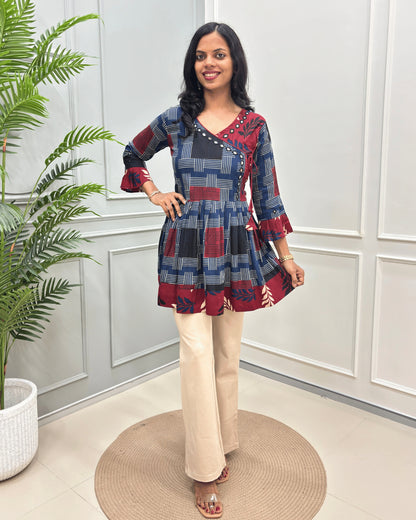Blue Women's Ajrakh Vegetable dyed plant based Cotton Short-Kurti with three-quarter frill sleeves, and waist to bottom,  paired with beige flared pant , standing on a round woven rug in front of a white paneled wall with a potted plant on the left.