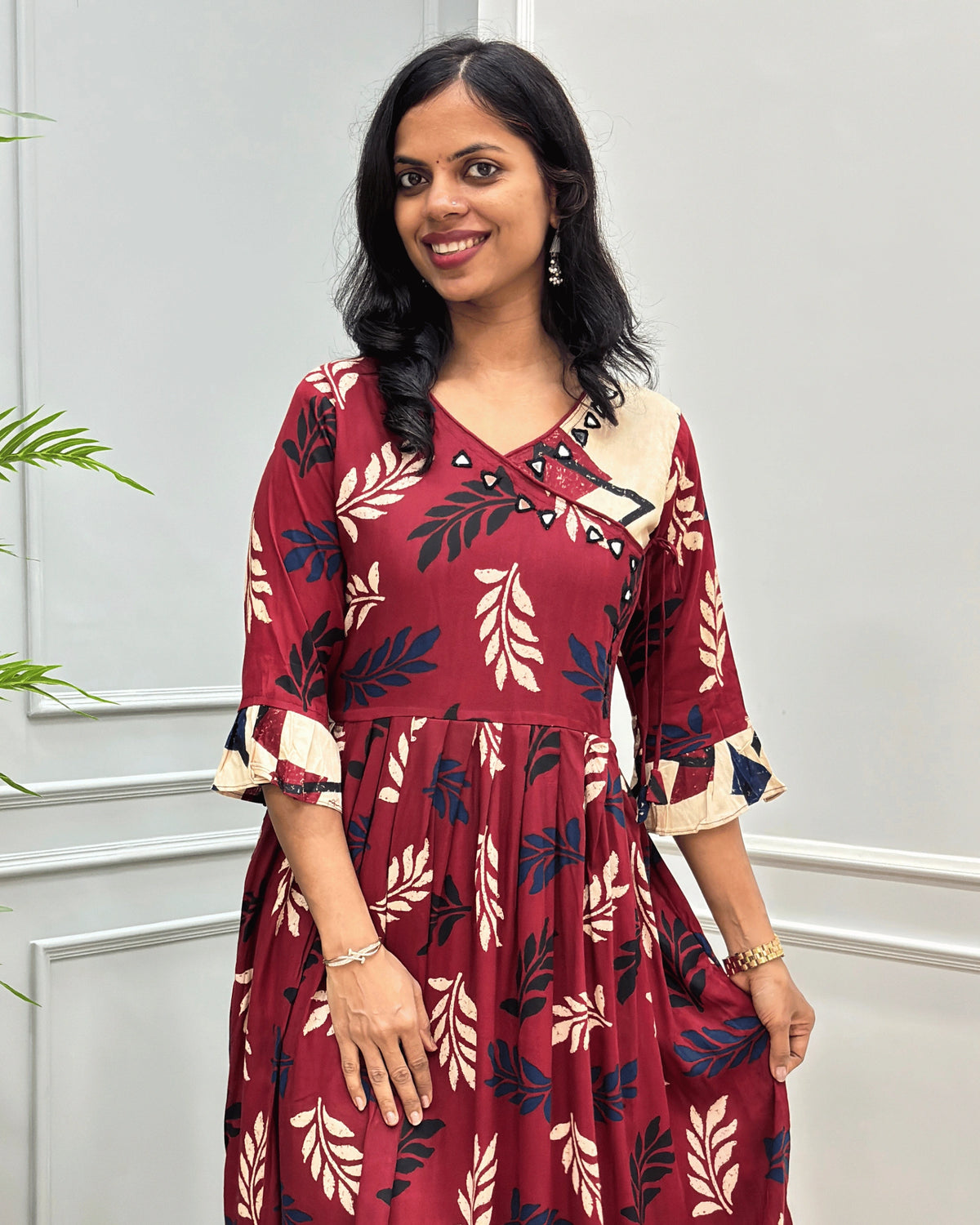 Close up of Red plant based cotton long kurti with navy, and beige leaf-Print , Angrakha pattern with lace and hand-mirror work on V-shaped neck ,  ruffled sleeves, standing against a light gray paneled wall.