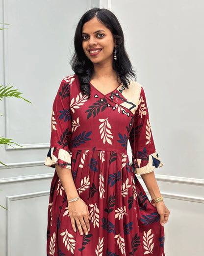 Close up of Red plant based cotton long kurti with navy, and beige leaf-Print , Angrakha pattern with lace and hand-mirror work on V-shaped neck ,  ruffled sleeves, standing against a light gray paneled wall.