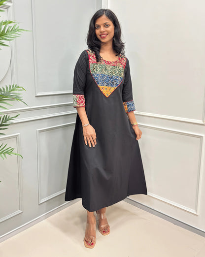 Black women's Ajrakh Vegetable dyed plant based cotton long kurti with a heavily embroidered multicolor Mashru yoke, standing in Front of white paneled room , on left side plotted plant .