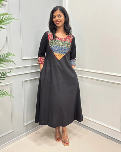 Front view of Black women's Ajrakh Vegetable dyed plant based cotton long kurti with a heavily embroidered multicolor Mashru yoke on front chest & sleeves cuffs along with  pocket, with red piepin, standing in Front of white paneled room , on left side plotted plant .
