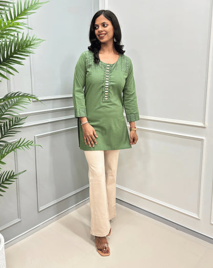 Teal-Green  women's Ajrakh Vegetable Dyed plant based Cotton Short-kurti with white piping and Hand-mirror work look a like button details, paired with beige Flared pants, standing in a room with a light gray paneled wall and a green plant on the left.