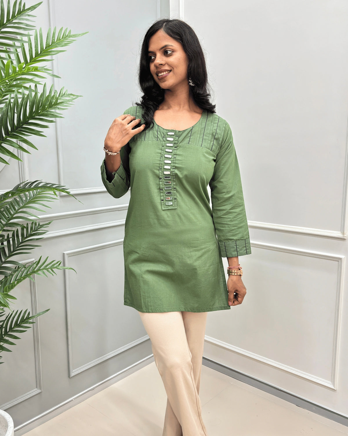 Front view of Navy-Blue women's Ajrakh Vegetable Dyed plant based Cotton Short-kurti with Black piping and Hand-mirror work look a like button details, Round neck paired with beige Flared pants, standing in a room with a light gray paneled wall and a green plant on the left.