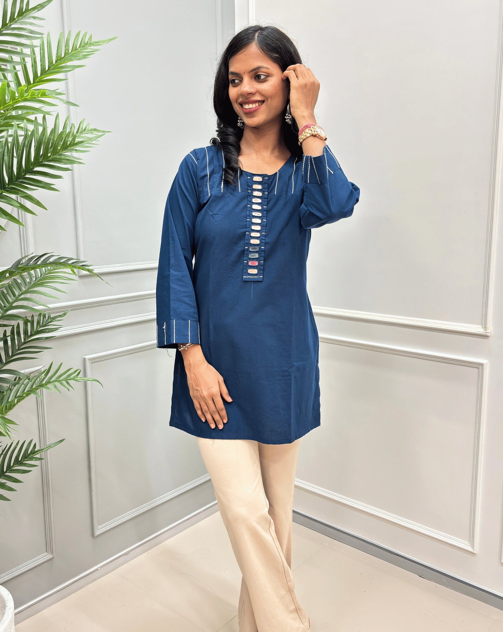 Front view of Navy-Blue women's Ajrakh Vegetable Dyed plant based Cotton Short-kurti with Beige piping and Hand-mirror work look a like button details, Round neck paired with beige Flared pants, standing in a room with a light gray paneled wall and a green plant on the left.