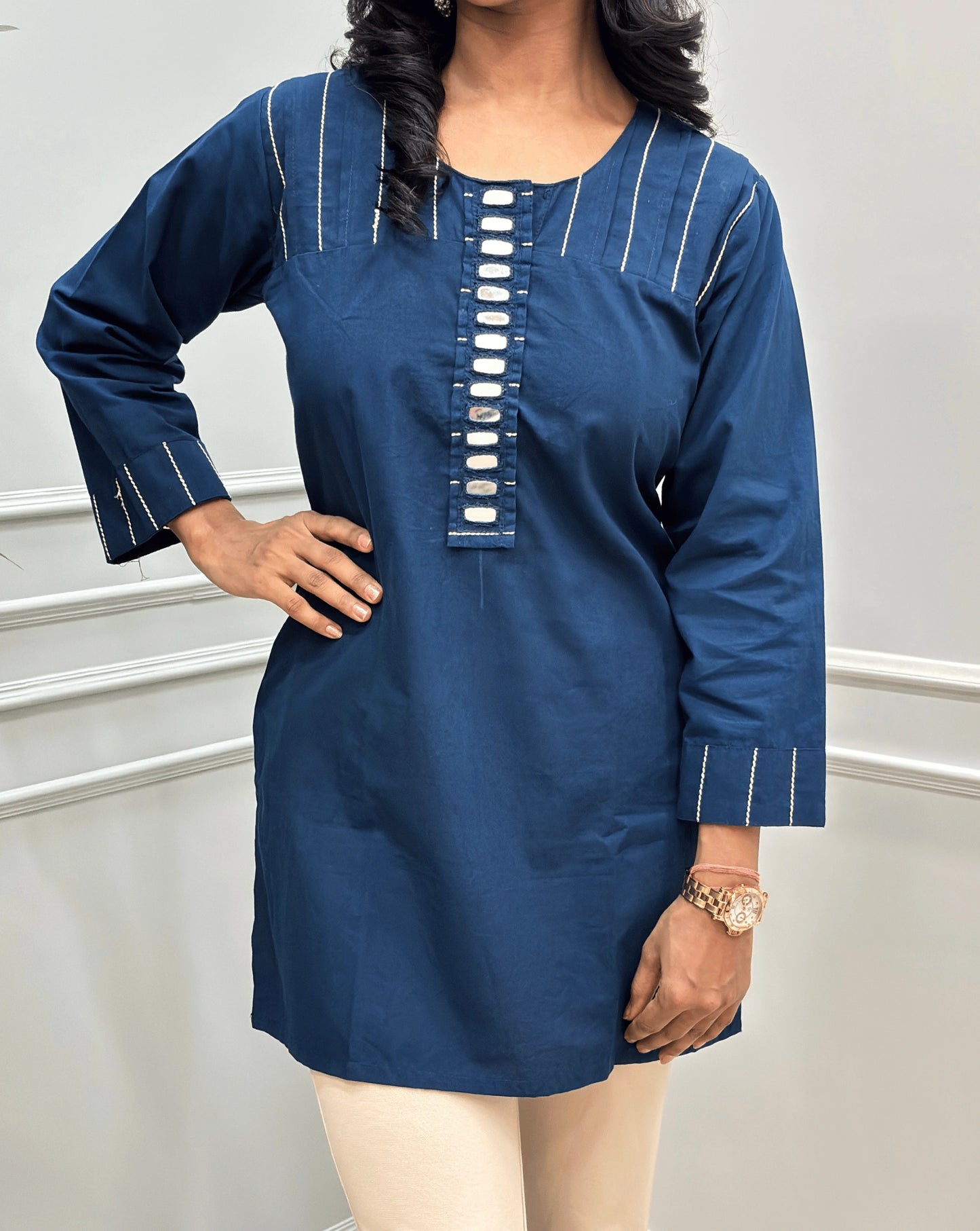 Close-up of Navy-Blue women's Ajrakh Vegetable Dyed plant based Cotton Short-kurti with Beige piping and Hand-mirror work look a like button details, Round neck & Full Sleeves  paired with beige Flared pants, standing in a room with a light gray paneled wall and a green plant on the left.