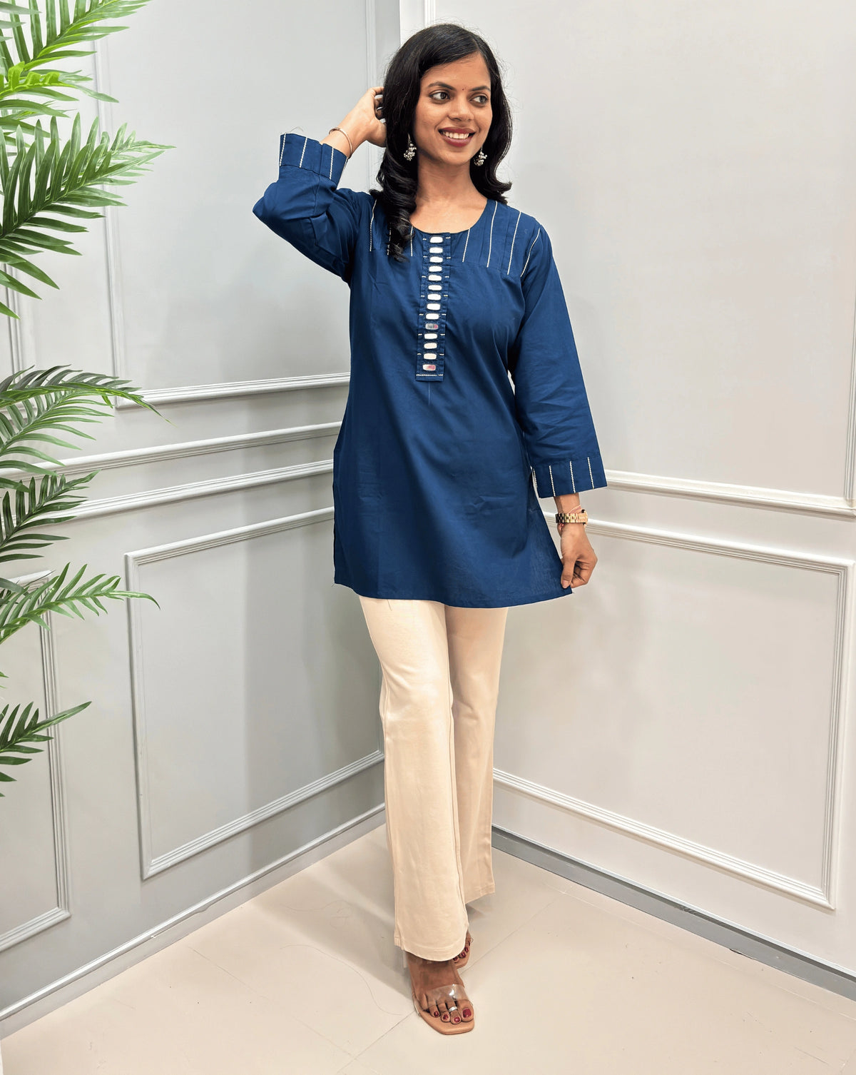 Navy-Blue women's Ajrakh Vegetable Dyed plant based Cotton Short-kurti with white piping and Hand-mirror work look a like button details, paired with beige Flared pants, standing in a room with a light gray paneled wall and a green plant on the left.
