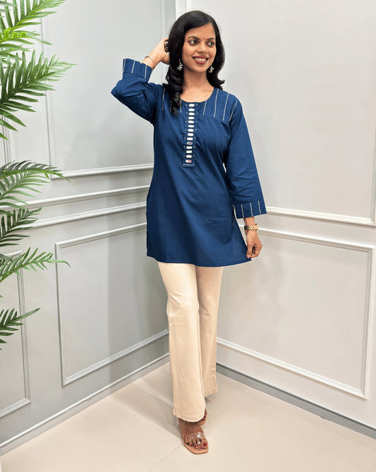 Navy-Blue women's Ajrakh Vegetable Dyed plant based Cotton Short-kurti with white piping and Hand-mirror work look a like button details, paired with beige Flared pants, standing in a room with a light gray paneled wall and a green plant on the left.