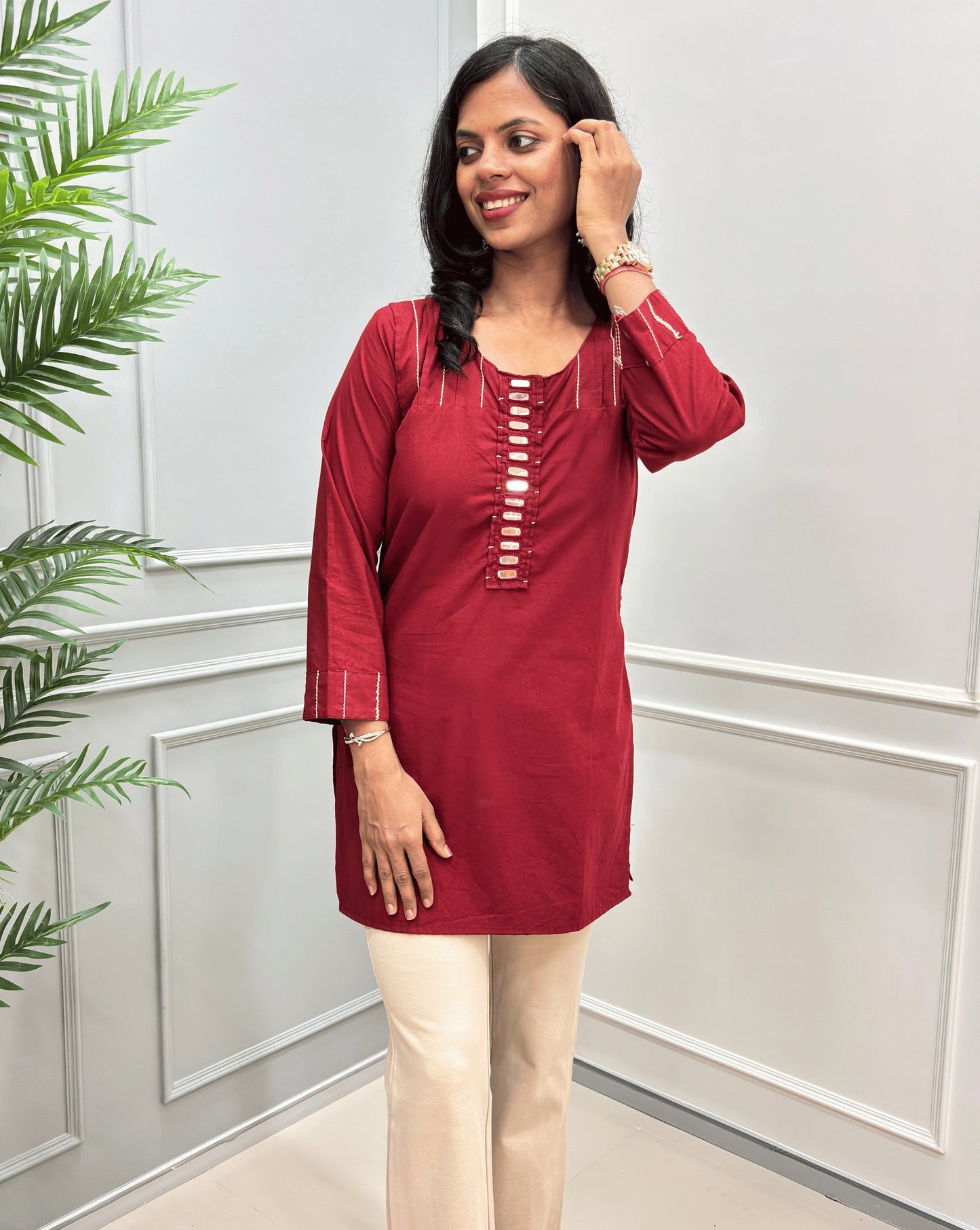 Front view of Deep Red women's Ajrakh Vegetable Dyed plant based Cotton Short-kurti with Beige piping and Hand-mirror work look a like button details, Round neck paired with beige Flared pants, standing in a room with a light gray paneled wall and a green plant on the left.